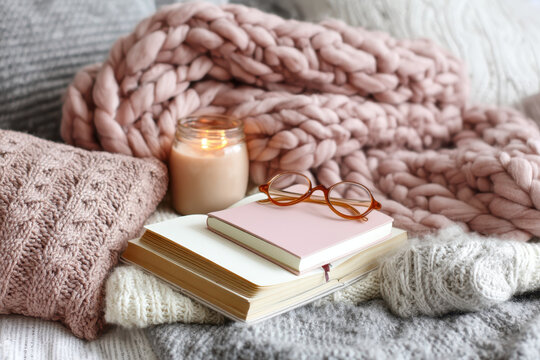 Cozy scene with pink knitted blanket, candle, and pair of glasses on stack of books, creating warm and inviting atmosphere