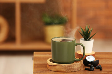 Cup of hot coffee and modern earphones on wooden table in room