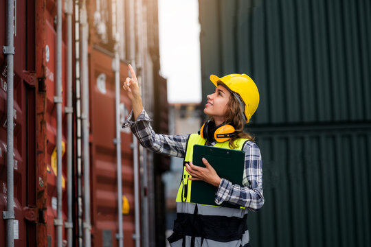 A woman wearing a yellow helmet and a safety vest is pointing at something on a container.