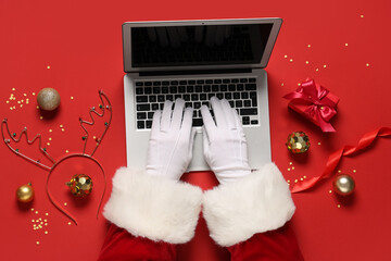 Santa Claus hands with modern laptop, Christmas decorations and gift box on red background, closeup