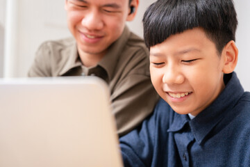 Close up of two Asian sibling study online learning, teenage brother teach younger boy with smile at home, looking at computer laptop, have fun, happiness and joy. Education and technology e-learning