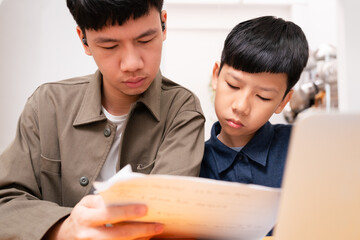 Two Asian sibling study together, reading, looking at lesson note with concentration. Teenage brother teach younger boy at home. Homeschool education, e-learning technology.