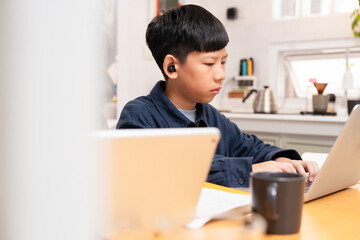 Smart looking Asian boy study online learning, type on computer laptop, listen with earbud with concentration and focus at home in white cozy kitchen. Homeschool education, e-learning technology