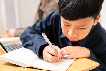 Close up of smart looking Asian boy write note on notebook, study online learning with concentration and focus at home on wooden table in white cozy room. Homeschool education, e-learning technology