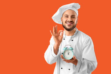 Young happy male chef with alarm clock showing OK gesture on orange background