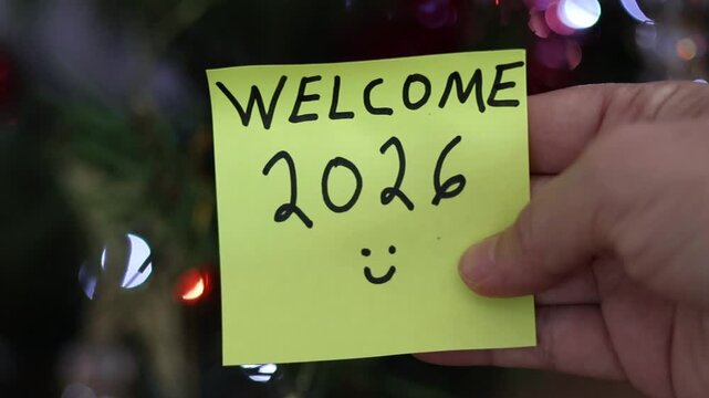 Un primer plano de la mano de una nota de color amarillo  con la palabra bienvenido 2026 en un fondo de luces de colores. Comienzo de a&ntilde;o