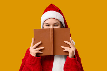Young woman in Santa hat with book on yellow background. Christmas concept