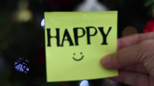 Una toma de cerca de una mano de mujer con una nota adhesiva de color amarillo con la palabra feliz en movimiento en un fondo de luces de colores