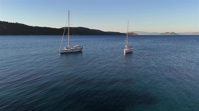 Greece,Kalamos Island,Aerial view forward towards Kastos island passing in between the two white sailboats anchored. Calm seas and crystal clear waters on a sunny day.