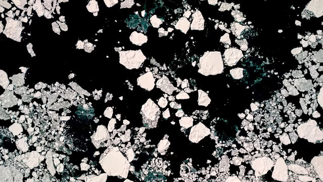 Abstract overhead shot of large and small chunks of white ice floating on dark, open water, resembling broken sea ice or floes