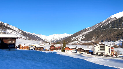A wide view of a snowy alpine village nestled in a valley, flanked by dark, forested slopes and distant snow-capped mountains under clear sky
