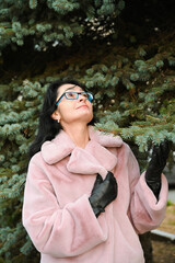 Happy russian 50 year old woman in glasses, winter coat and gloves looking and enjoying the fir tree