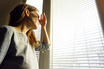 A woman raises her hand to block intense daylight streaming through window blinds during a severe headache flare. Symbolizes light sensitivity, migraine trigger response, and chronic pain struggle. 