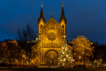 Fototapeta premium Illuminated Christmas Tree in Karlín, Prague, with the Church of St Cyril and Methodius During Winter Holiday Evening