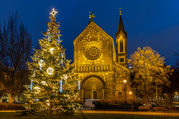 Fototapeta premium Illuminated Christmas Tree in Karlín, Prague, with the Church of St Cyril and Methodius During Winter Holiday Evening