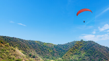 Outdoor scene featuring a paraglider soaring through a clear blue sky over lush green hills. Natural background