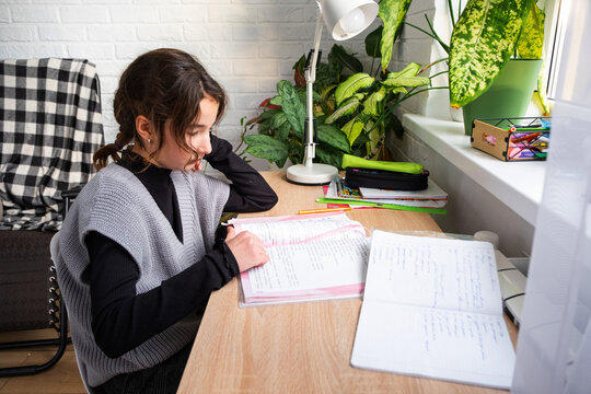 A schoolgirl girl is doing her homework from school at the table, writing in a notebook - Powered by Adobe