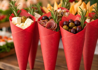 Gourmet charcuterie cones filled with assorted cheeses, olives, cured meats and rosemary sprigs, wrapped in elegant red paper, served at a holiday market.