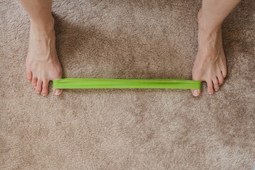 Woman doing foot rehabilitation exercise with elastic band, closeup photo. Foot stretching with resistance band on soft carpet for home physical therapy