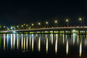 Illuminated bridge in Hue at night featuring starburst streetlights, red flags, and golden reflections on the dark river water.
