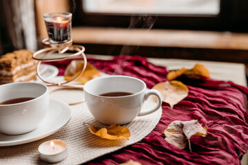 A cozy still life with black tea in white cups on a burgundy cloth, candles flickering near a slice of cake.