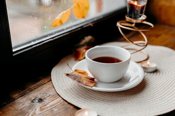A cup of black tea by a rainy window on a wooden sill, candlelight glowing nearby for a soft intimate mood.