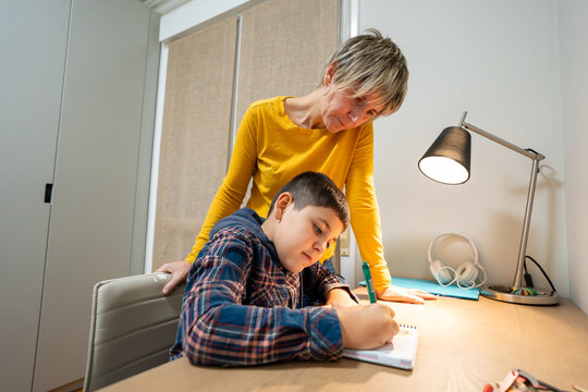 Mother watching son writing in notebook, assisting with home education and studying at a desk with a lamp