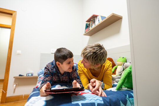 Loving mother and child with down syndrome lying on bed, looking at each other while using a digital tablet
