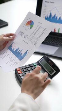 A tight close-up over-the-shoulder video of hands in a formal business jacket sleeve operating a modern calculator, cross-referencing crisp financial documents with a professional laptop graph on an