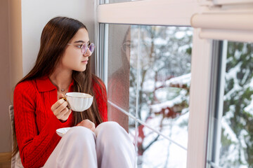Girl sipping tea and looking out snowy window. Teen girl in red sweater and glasses holding a teacup, peacefully gazing out the window at a snowy winter landscape.