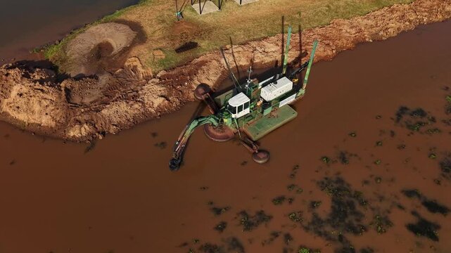 Aerial orbit over amphibious dredging machine operating on muddy river shoreline during afternoon with intense sun rays.