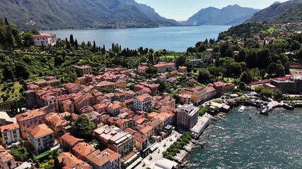High-angle drone glides over Bellagio peninsula on Lake Como, Italy, pastel-roofed buildings along the lakefront, cypress-lined promenade, and lush gardens merging into water, with Lombardy Alps