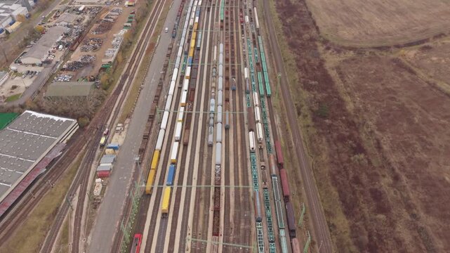 A wide aerial view of a large railway yard filled with freight trains and cargo wagons arranged on multiple parallel tracks. A green yellow passenger train passes through.