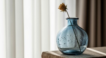 A blue glass vase with a dried rose on a stone table. Minimalist interior. A wilted yellow flower in a bubble glass bottle against a backdrop of white curtains.