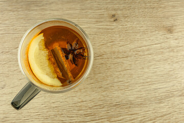 A clear double-walled glass filled with hot tea with oriental spices, cinnamon sticks, hibiscus, cloves, orange, star anise, a warming drink in a wooden table