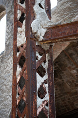 Close-up of a rusted metallic lattice column and beam junction in an abandoned factory, showing exposed rivets, heavy corrosion, and broken cement coating.