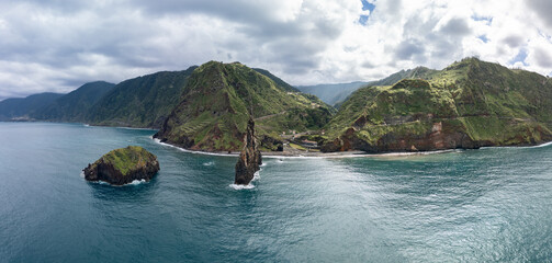 Panoramic drone view of Ilheus da Ribeira da Janela on Madeira island, showing volcanic sea stacks, terraced green cliffs and Atlantic waves
