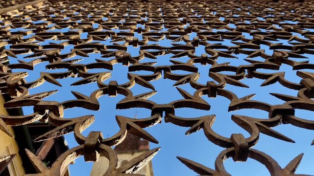  antique wrought-iron fence or gate section, featuring a repeated pattern of rustic, curved arrow-like shapes. The rusted ironwork is silhouetted against a bright blue sky