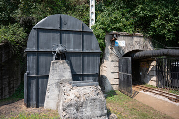 Exterior of a giant, historical black centrifugal fan by Victor Gruber next to the gated, abandoned coal mine tunnel entrance, surrounded by greenery.