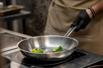 Professional chef in black gloves cooking fresh vegetables and seafood in a stainless steel pan on an induction stove, steam rising in a restaurant kitchen setting