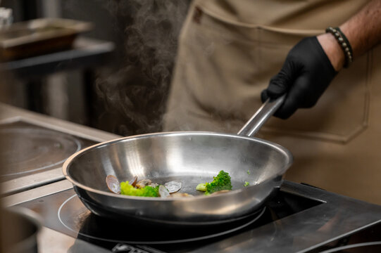 Professional chef in black gloves cooking fresh vegetables and seafood in a stainless steel pan on an induction stove, steam rising in a restaurant kitchen setting