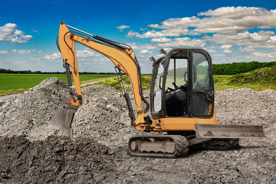 Mini excavator digs into a large pile of dirt at a construction site under a bright blue sky with fluffy clouds and green fields nearby - Powered by Adobe
