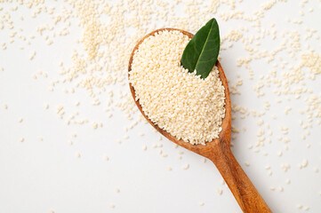 Close-up of white sesame seeds in wooden spoon with green leaves on light white background. Healthy organic seeds, cooking ingredient. Sesamum indicum. Top view flat lay
