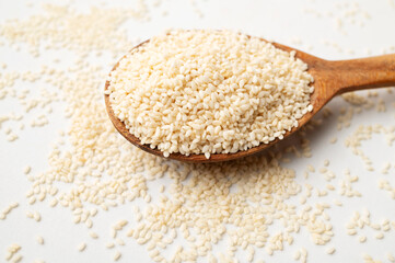 Close-up of white sesame seeds in wooden spoon with green leaves on light white background. Healthy organic seeds, cooking ingredient. Sesamum indicum. Top view flat lay