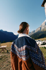 Fototapeta premium Woman in a patterned sweater stands with her back to camera in the mountains outdoors, overlooking a quiet countryside landscape under clear sky, enjoying autumn light and peaceful nature.