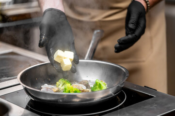 Close-up of a professional chef in black gloves adding butter cubes to a frying pan with fresh broccoli and other ingredients on an electric stovetop. Steam rises as the food cooks