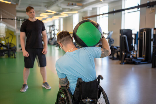 Man in wheelchair training with medicine ball do bend stretch, head supported neck resistance for seated core stability. Therapeutic recovery exercise, inclusive strength development, adaptive sport
