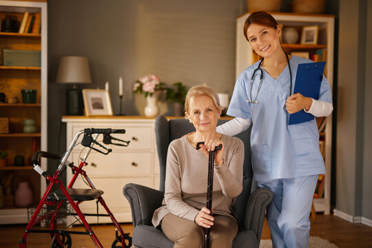 A friendly female healthcare worker in light blue scrubs visits a senior woman at her home. The woman sits in an armchair holding a cane, with her walker nearby.