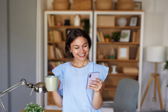 A woman in a light blue top smiles as she holds a cup of coffee in one hand and looks at her smartphone with the other. - Powered by Adobe