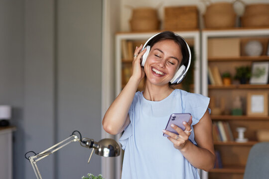 A young woman is smiling and enjoying music through headphones while holding a smartphone in her home office. The space features bright lighting and neatly arranged shelves.